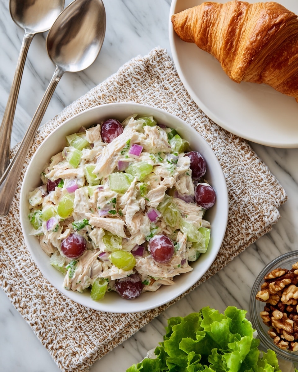 A white bowl filled with a creamy salad that has three main layers: the base is shredded light beige chicken mixed with white mayonnaise dressing, scattered with green celery pieces, red onion chunks, and halved red grapes. Around the bowl, there is a bottom left cluster of fresh green leaf lettuce, to the bottom right a small glass bowl with chopped walnuts, and on the top right edge, part of a white plate with a golden-brown flaky croissant, all set on a white marbled textured surface. A pair of metal spoons rest slightly behind the white salad bowl. Photo taken with an iphone --ar 4:5 --v 7