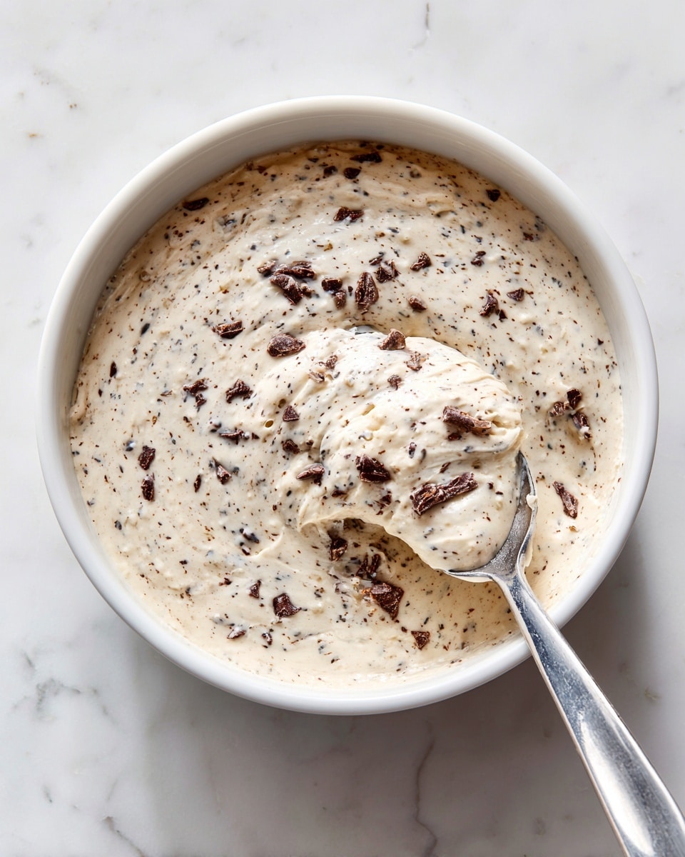 A close-up view of a single-layer creamy dish with a light beige color and a soft, slightly grainy texture, dotted with small, dark brown chocolate chips spread unevenly throughout, filling a white bowl with small handles on both sides. A silver spoon is sticking into the right side of the dish, partially covered by the creamy mixture. The bowl is placed on a white marbled surface with subtle gray veins. photo taken with an iphone --ar 4:5 --v 7