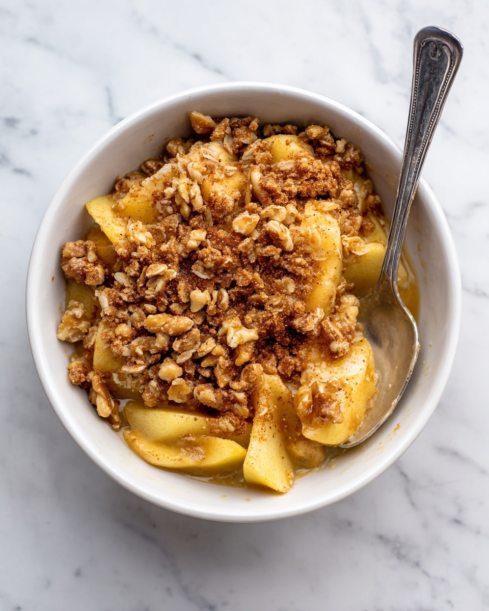 A white bowl filled with a layered apple crisp dessert sits on a white marbled surface. The base layer has soft, cooked apple slices in light golden yellow with a slightly translucent texture. On top, there is a crunchy granola topping made of oats and nuts that are golden brown and slightly clumped together, sprinkled evenly across the apples. A small spoon rests inside the bowl on the right side. The lighting is soft and natural, highlighting the warm tones of the apples and the crisp texture of the granola photo taken with an iphone --ar 4:5 --v 7