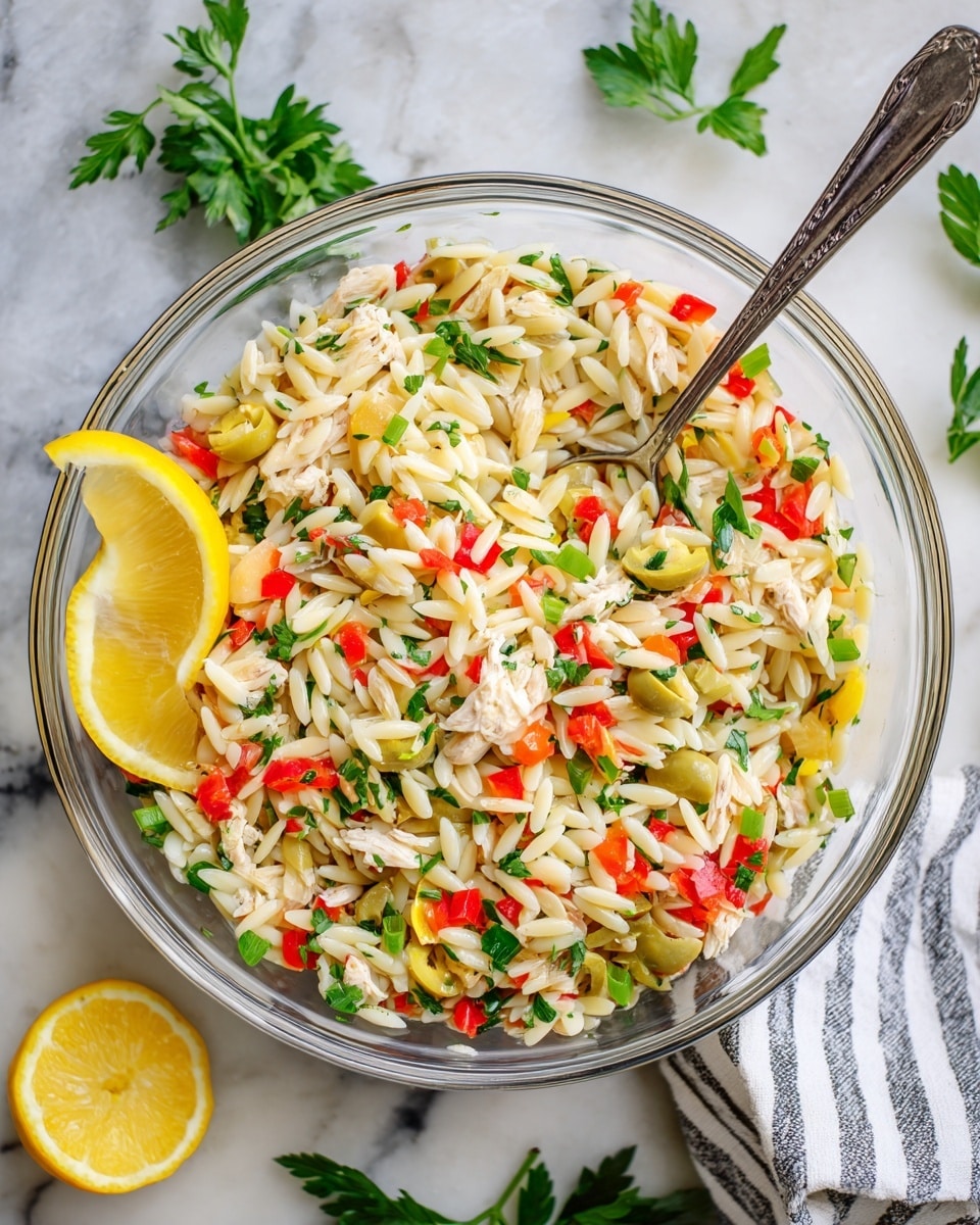A clear glass bowl filled with a colorful mixed salad resting on a white marbled surface; the salad has four main layers: a base layer of small white rice-shaped pasta, mixed with small chunks of light beige shredded chicken; the second layer consists of diced red bell peppers and yellow bell peppers scattered evenly; the third layer has small pieces of green olives and chopped green herbs mixed throughout; on top there are fresh parsley leaves for garnish and three lemon wedges placed on one side for decoration. A silver spoon is partially visible inside the bowl. photo taken with an iphone --ar 4:5 --v 7