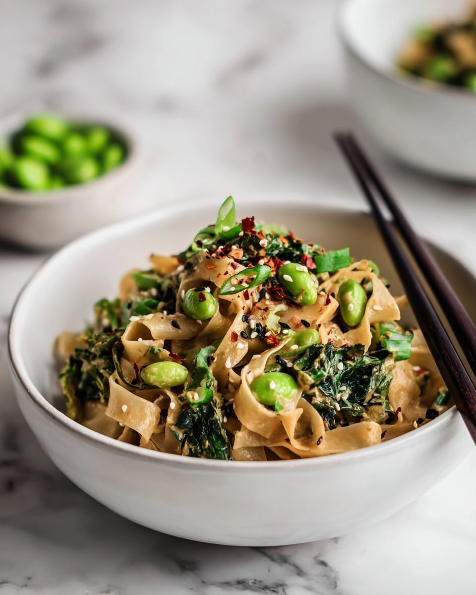 A white bowl filled with three main layers: at the base are wide, flat noodles with a light brown sauce, mixed with bright green edamame beans and dark green leafy vegetables. On top, there are scattered white and black sesame seeds along with red chili flakes, adding small dots of black, white, and red. Small green onion pieces are sprinkled throughout, giving fresh green highlights. Black chopsticks rest on the bowl's edge. The bowl sits on a wooden surface with a blurred white marbled background. photo taken with an iphone --ar 4:5 --v 7