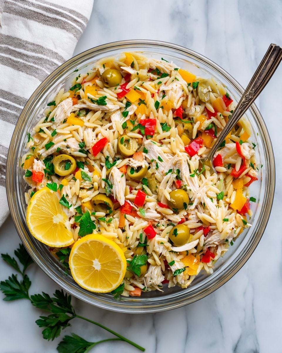 A clear glass bowl filled with a colorful orzo chicken pasta salad, showing small pasta grains mixed with shredded white chicken pieces, diced red and yellow bell peppers, sliced green olives, and chopped green herbs. Two lemon wedges with bright yellow peel sit on one edge, and fresh green parsley decorates the bottom. A silver spoon rests inside the bowl, partially visible on the right side, all placed on a white marbled surface with a white and gray striped cloth nearby. Photo taken with an iphone --ar 4:5 --v 7