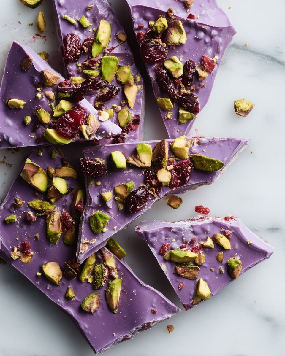 This image shows broken pieces of a thin, flat purple chocolate bar spread on a white marbled surface. The chocolate has a smooth texture and is topped with scattered chunks of bright green pistachios and dark red dried berries, which create a colorful contrast. The pieces are irregularly shaped with rough edges, and bits of chocolate, pistachios, and berries are scattered loosely around. The lighting highlights the glossy finish of the chocolate and the textures of the toppings. photo taken with an iphone --ar 4:5 --v 7