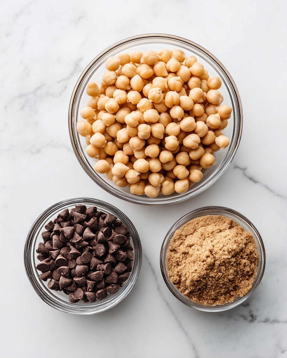A white marbled surface holds three clear glass and metal bowls arranged in a triangle, each filled with a single ingredient. The top bowl, round and clear glass, is full of round, pale beige chickpeas with a smooth texture. Below to the left, a metal measuring cup holds dark brown chocolate chips, small and shiny with a slightly rounded shape. To the right, another metal measuring cup contains a finely ground light brown powder, likely brown sugar, with a soft, crumbly texture. The bowls are neatly placed on the clean white marbled surface. Photo taken with an iphone --ar 4:5 --v 7