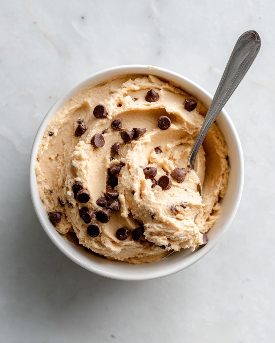 A white bowl filled with creamy, light beige dough with a soft and slightly grainy texture, mixed with scattered dark brown chocolate chips. The dough is thick and dolloped unevenly in the bowl with some small peaks and valleys visible, giving a fresh, homemade look. The bowl sits on a white marbled surface that adds a clean, bright touch to the image. photo taken with an iphone --ar 4:5 --v 7