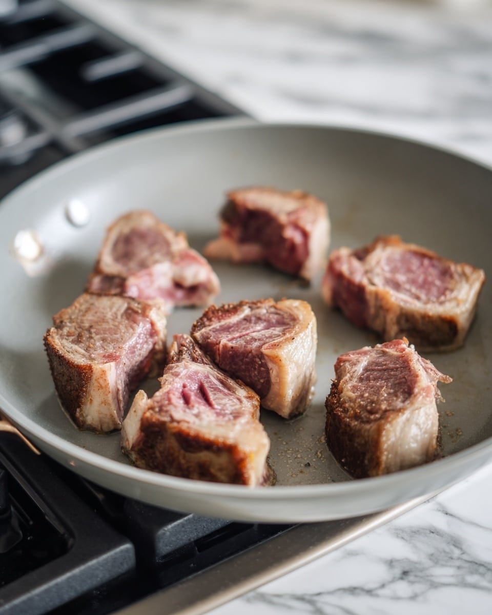 A close-up view of a light gray pan on a stove with several pieces of meat inside. There are six pieces in total, varying in size and shape. Each piece has a browned, cooked outer edge with a mix of pink and red inside, showing their rawness in the middle. The meat pieces have visible bones in the center and some fat around the edges. The pan surface is smooth and clean, and the background shows the black stove burners surrounded by white marbled texture. photo taken with an iphone --ar 4:5 --v 7