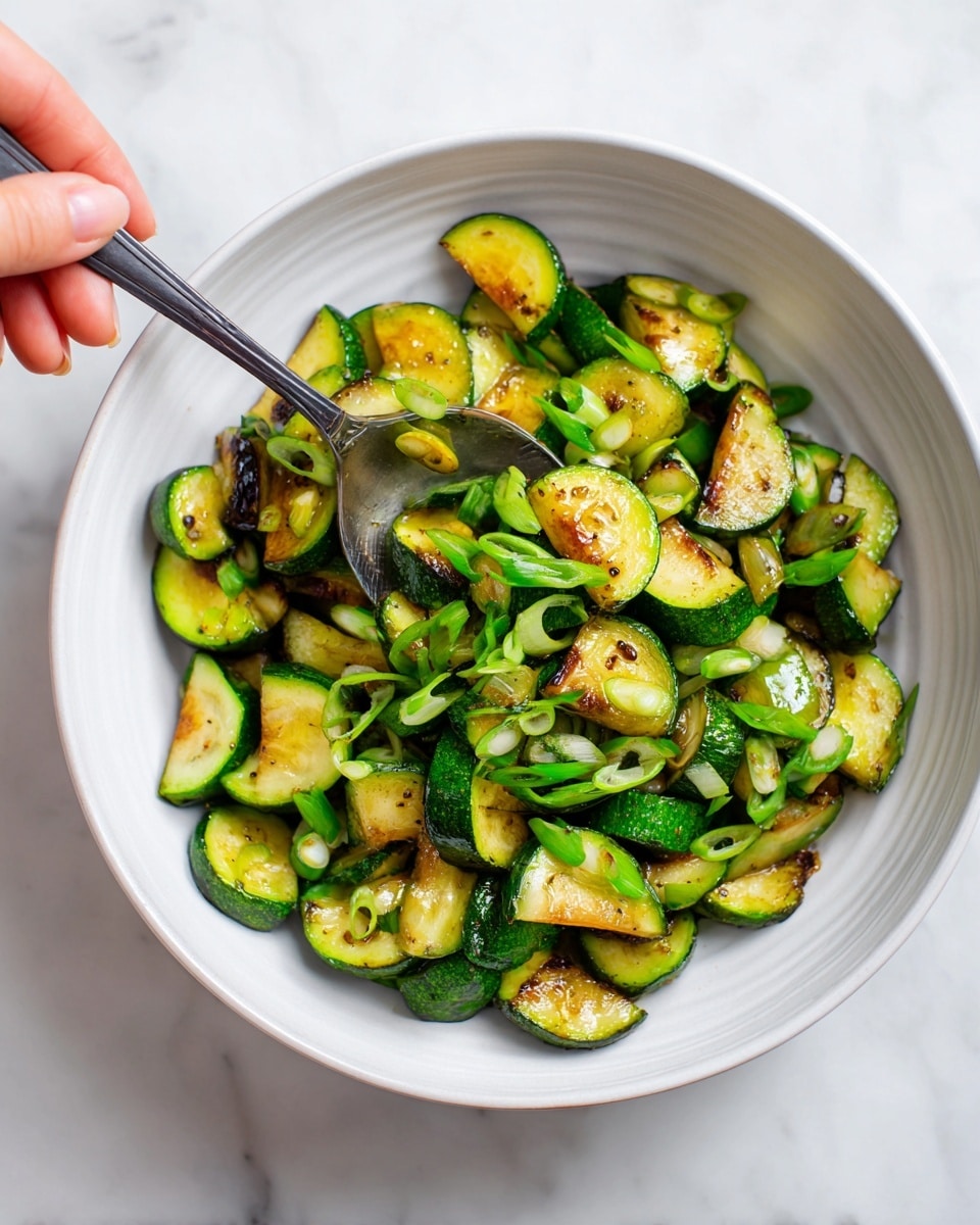 The image shows a white bowl filled with cooked chopped zucchini pieces that have a golden brown seared texture on some sides, giving them a slightly crispy look. Mixed in with the zucchini chunks are thin slices of bright green scallions scattered throughout, adding a fresh contrast. A woman’s hand is holding a silver spoon that is scooping up some of the zucchini and scallions from the bowl. The bowl sits on a white marbled textured surface that is softly lit. The colors are mostly green and yellow-brown with a shiny, cooked texture on the vegetables. photo taken with an iphone --ar 4:5 --v 7