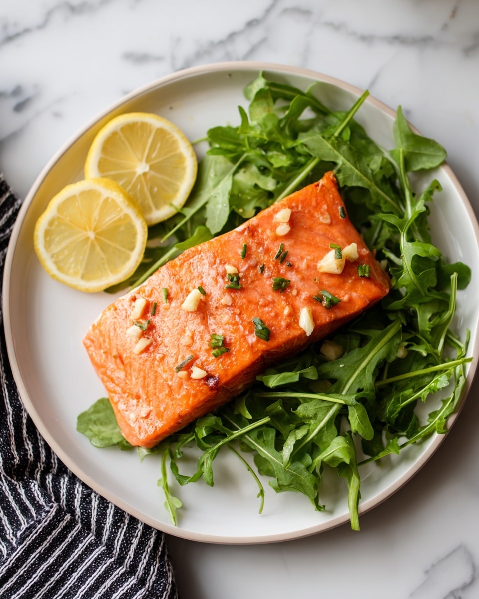 A single piece of cooked salmon with an orange-pink color sits on a bed of bright green arugula leaves, topped with small bits of garlic and herbs scattered evenly over its surface, showing a slightly crisp texture. To the left of the salmon are two lemon wedges with a shiny, juicy look. All the food rests on a white plate with a subtle rim, placed on a white marbled surface with part of a black-and-white striped cloth napkin visible on the lower left. photo taken with an iphone --ar 4:5 --v 7