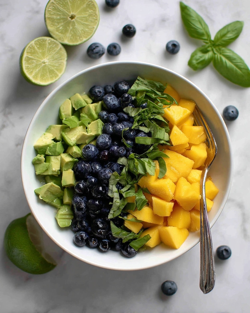 A white bowl holds a colorful fruit salad with three visible layers: the bottom layer is small green avocado pieces, the middle layer is bright yellow mango cubes, and the top layer has dark blue blueberries mixed with chopped green basil leaves. A silver fork rests inside the bowl on the right side. On the left edge of the bowl, there is a lime wedge with a light green color and visible segments. Around the bowl, on a white marbled surface, there are scattered blueberries, lime slices, and some basil leaves for decoration. photo taken with an iphone --ar 4:5 --v 7