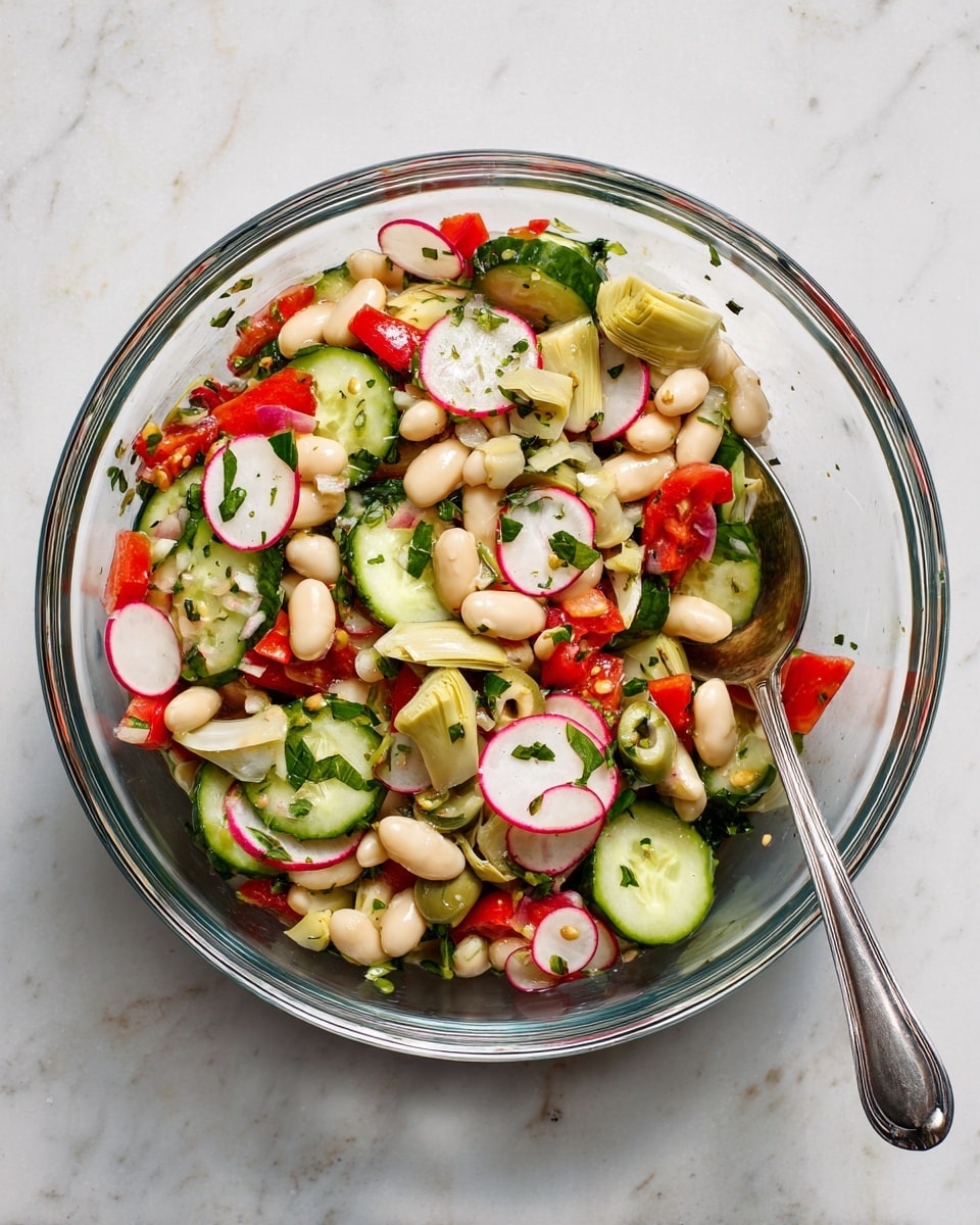 A clear glass bowl filled with fresh salad sits on a white marbled surface. The salad has many layers including crisp light green cucumber slices, red and white radish slices, and small white beans. There are small pieces of light yellow artichoke hearts mixed in with chopped red bell peppers and halved cherry tomatoes. Bright green chopped herbs and sliced green olives with pimento are scattered all over, adding color contrast. A silver spoon rests inside the bowl, partially covered by the salad. The textures look fresh and crunchy, and the colors are bright and natural photo taken with an iphone --ar 4:5 --v 7