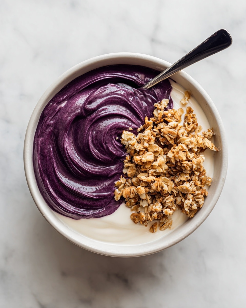 A white bowl holds two main layers: the bottom layer is smooth and white, likely yogurt, and the top layer is a thick, deep purple smoothie spread on one side. On top of the purple layer, there is a generous pile of light brown granola with visible oat clusters. A spoon with a black handle is scooping from the purple layer, creating a swirl of dark purple color blending into the white layer. The bowl sits on a white marbled surface. photo taken with an iphone --ar 4:5 --v 7