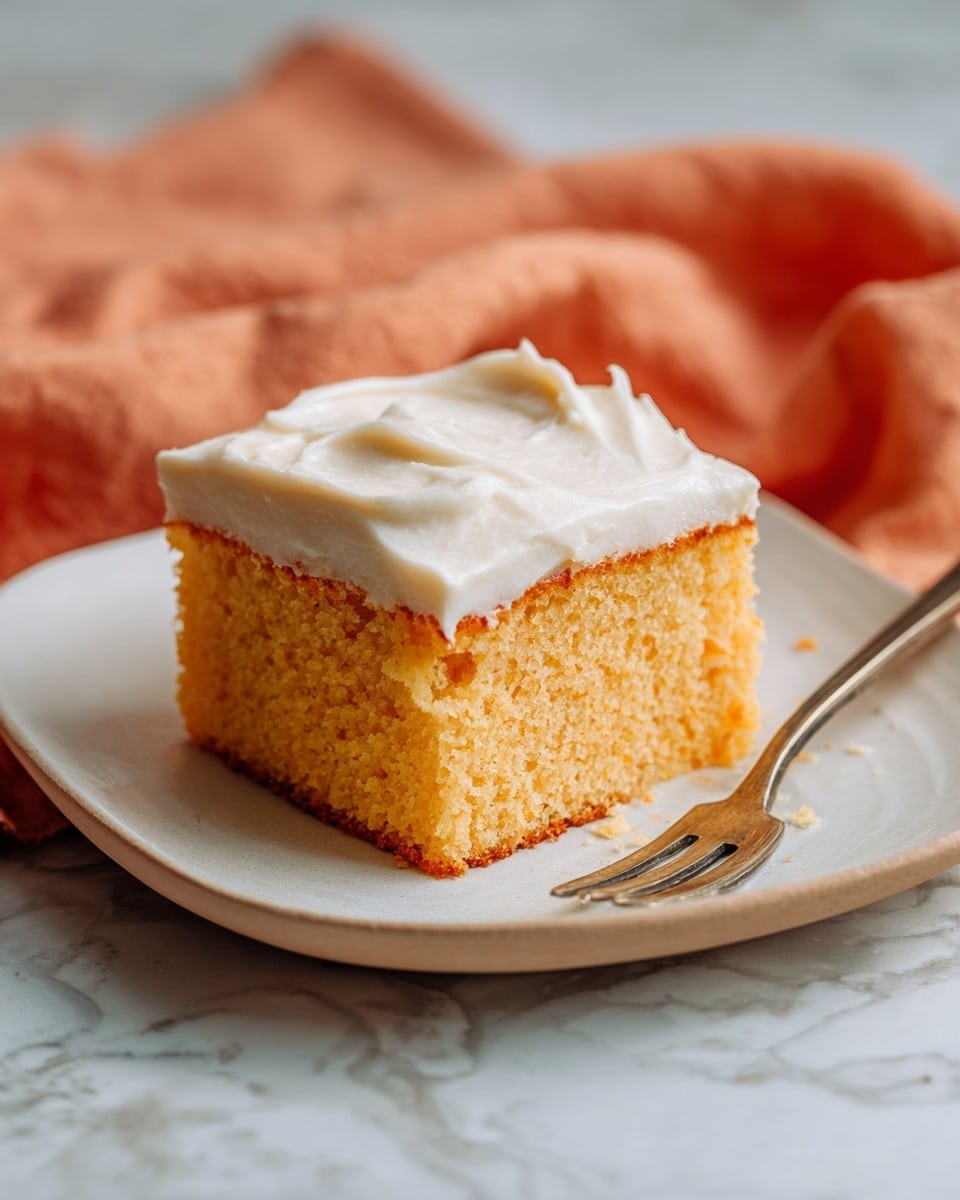 A thick square piece of moist orange-brown cake sits in the center of a white plate with a slightly scalloped edge, topped with a generous layer of smooth, creamy white frosting that has gentle swirls. The cake shows a soft, spongy texture with small air holes visible throughout. A silver fork rests beside the cake on the plate. The background has a white marbled texture with a soft orange cloth slightly visible on the side. Photo taken with an iphone --ar 4:5 --v 7