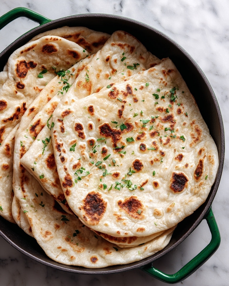 The image shows a close-up of four golden brown naan breads with uneven char marks, placed overlapping inside a dark pan with green handles. The naan has a soft, slightly puffy texture with some bubbly, browned spots and is sprinkled with small green herb leaves scattered on top. The pan rests on a white marbled surface. photo taken with an iphone --ar 4:5 --v 7