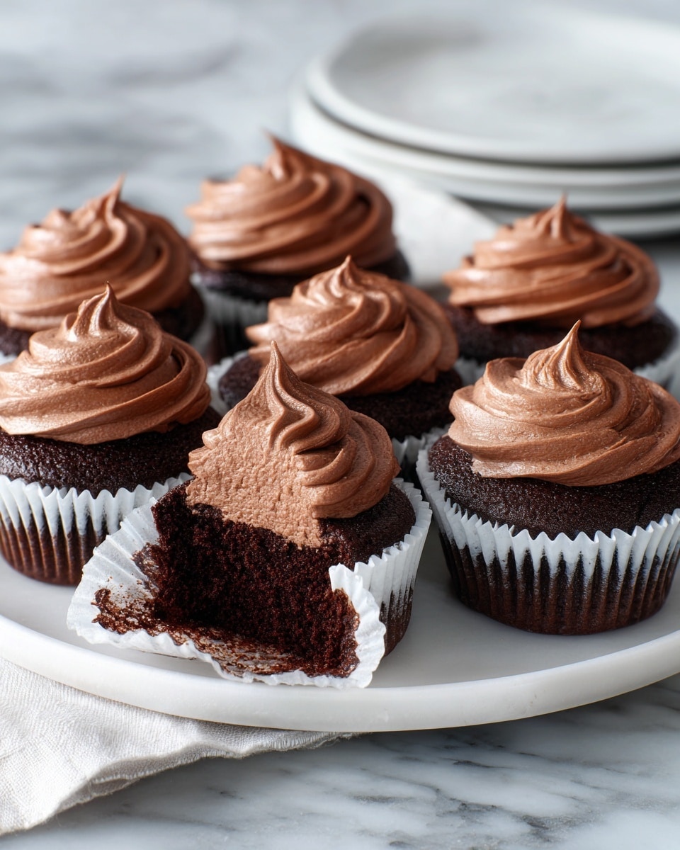 A white plate holds six chocolate cupcakes in white paper liners, each topped with a swirl of smooth, light brown chocolate frosting. One cupcake in the front is opened, showing its dark, moist chocolate inside with a thick layer of frosting on top. The opened cupcake’s silver liner is partially peeled back, resting on the plate. The cupcakes are arranged in a casual circle, and the background features a soft cream cloth and a white marbled surface. photo taken with an iphone --ar 4:5 --v 7