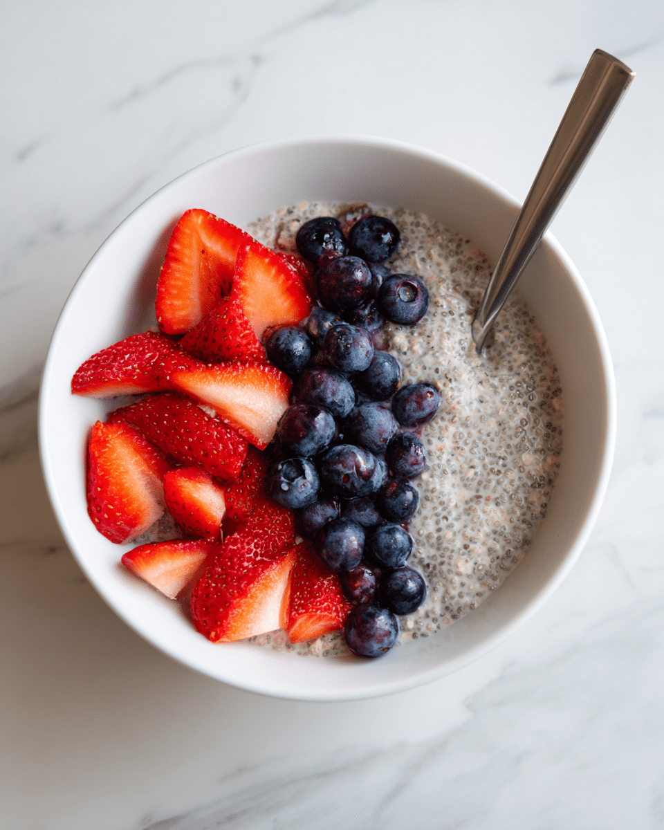 A white bowl filled with a creamy mixture of oats and chia seeds that looks soft and slightly thick, covering most of the bowl. On the right side of the bowl, there are fresh sliced strawberries and whole blueberries that add bright red and deep blue colors, with a shiny silver spoon resting inside the bowl next to the fruit. The bowl is placed on a white marbled surface. Photo taken with an iphone --ar 4:5 --v 7