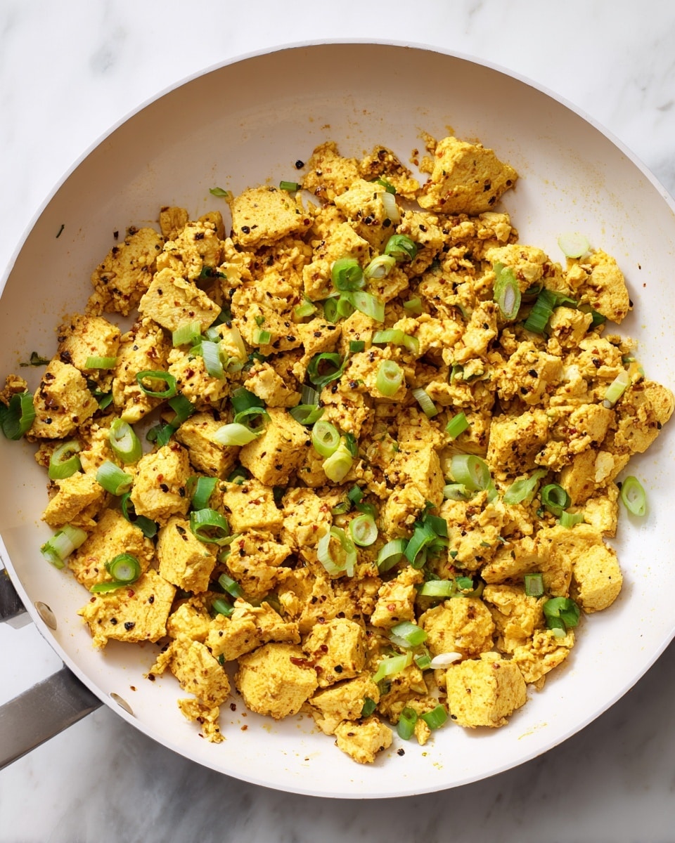 The image shows a white scalloped-edged plate holding a morning meal with three parts. On the right half, there is a large pile of soft, crumbly scrambled yellow tofu with bits of chopped green onion sprinkled on top. On the left side, there are two slices of golden-brown toasted bread leaning on each other, showing a crispy texture. In the bottom left corner of the plate, there are five lightly roasted bright red cherry tomatoes with some slight browning from roasting. All of this is on a white marbled background. photo taken with an iphone --ar 4:5 --v 7