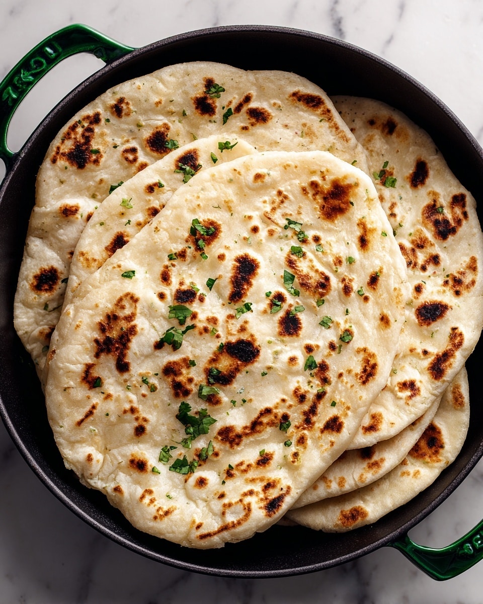 The image shows four flatbreads with a slightly uneven, golden-brown surface, each with patches of darker brown spots from cooking. They are layered slightly overlapping each other in a black pan. The flatbreads have a soft, fluffy texture and are sprinkled with small green herb pieces, adding contrast to the warm tones of the bread. The overall look is rustic and inviting, set against a white marbled surface. photo taken with an iphone --ar 4:5 --v 7