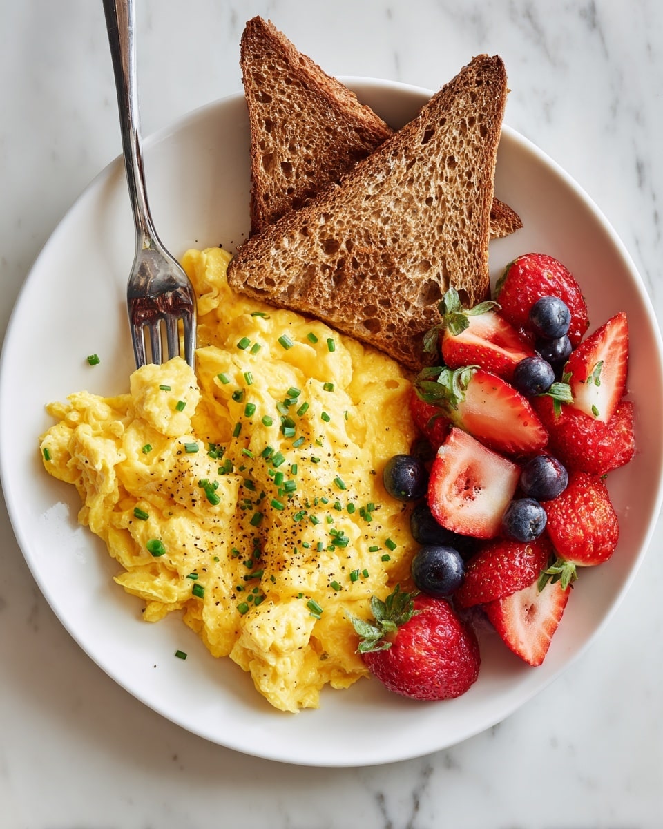 A white plate sits on a white marbled surface, holding a breakfast of three main parts: a pile of fluffy yellow scrambled eggs sprinkled with small green herbs and black pepper near the center, two triangular pieces of golden brown toast placed overlapping at the top of the plate, and a small cluster of fresh fruit on the right side including several bright red strawberries and round deep blue blueberries. A silver fork with an ornate handle rests on the plate's left edge, partly touching the scrambled eggs. Photo taken with an iphone --ar 4:5 --v 7