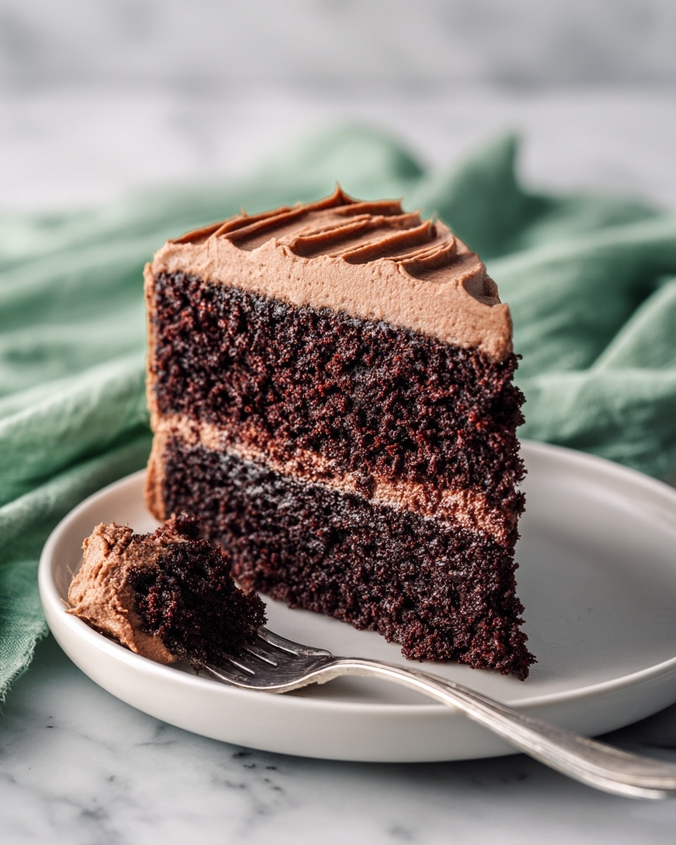 A two-layer dark chocolate cake slice sits on a white plate, each layer separated and covered with a thick creamy light brown frosting. The frosting has smooth swirls and ridges on the sides of the cake, adding texture and depth. A silver fork rests on the plate near the cake, with a small piece of the cake and frosting on its tines. The background has a soft white marbled texture, and a green cloth napkin is partly visible beside the plate. Photo taken with an iphone --ar 4:5 --v 7