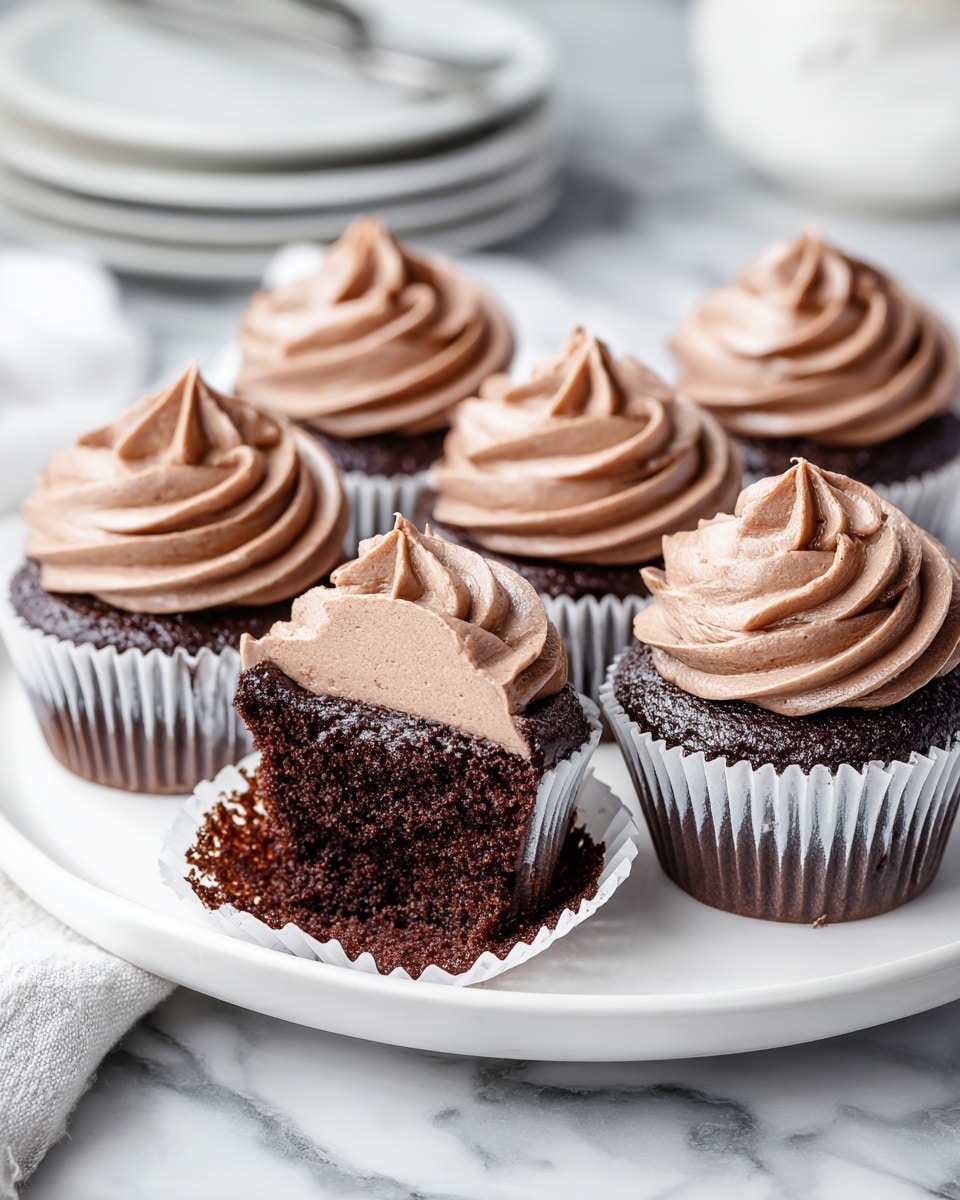 A white round plate holds six chocolate cupcakes with dark brown cake bases in white paper liners. Each cupcake is topped with a smooth swirl of rich, creamy chocolate frosting that has a glossy, slightly thick texture. One cupcake at the front center is partially unwrapped with silver foil peeled down and has a bite taken out of it, showing the moist, dense dark cake inside and a thick layer of the same chocolate frosting on top. The plate is placed on a soft beige cloth on a white marbled textured surface, with a blurred open laptop and another plate holding more cupcakes in the background. photo taken with an iphone --ar 4:5 --v 7