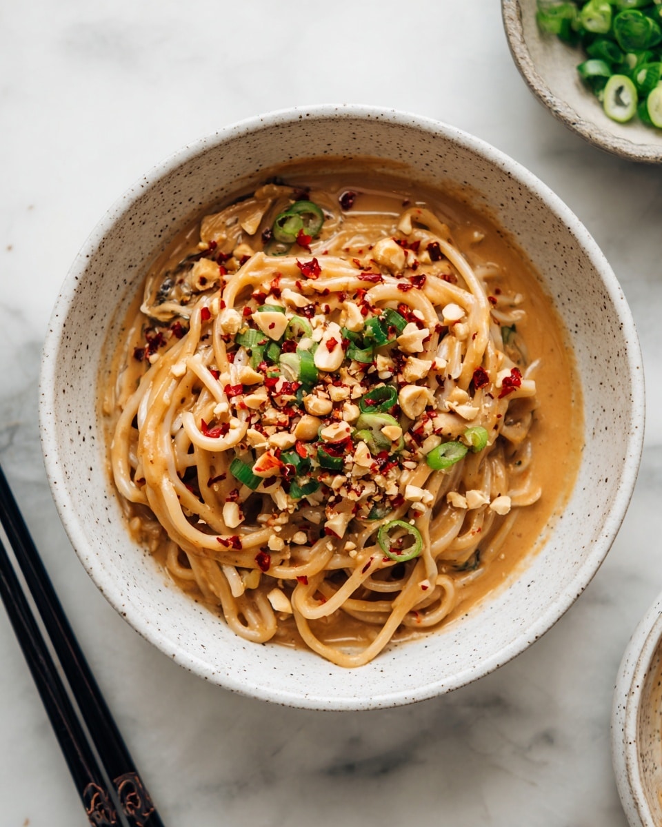 A close-up view of thick light brown noodles coated in a creamy sauce piled inside a round white bowl. The noodles are topped with crushed light tan peanuts, thinly sliced bright green scallions, and small red chili flakes sprinkled evenly across. Black chopsticks rest on the left edge, contrasted against the white marbled surface underneath. A small bowl with chopped green scallions is placed slightly out of focus in the top right corner. photo taken with an iphone --ar 4:5 --v 7