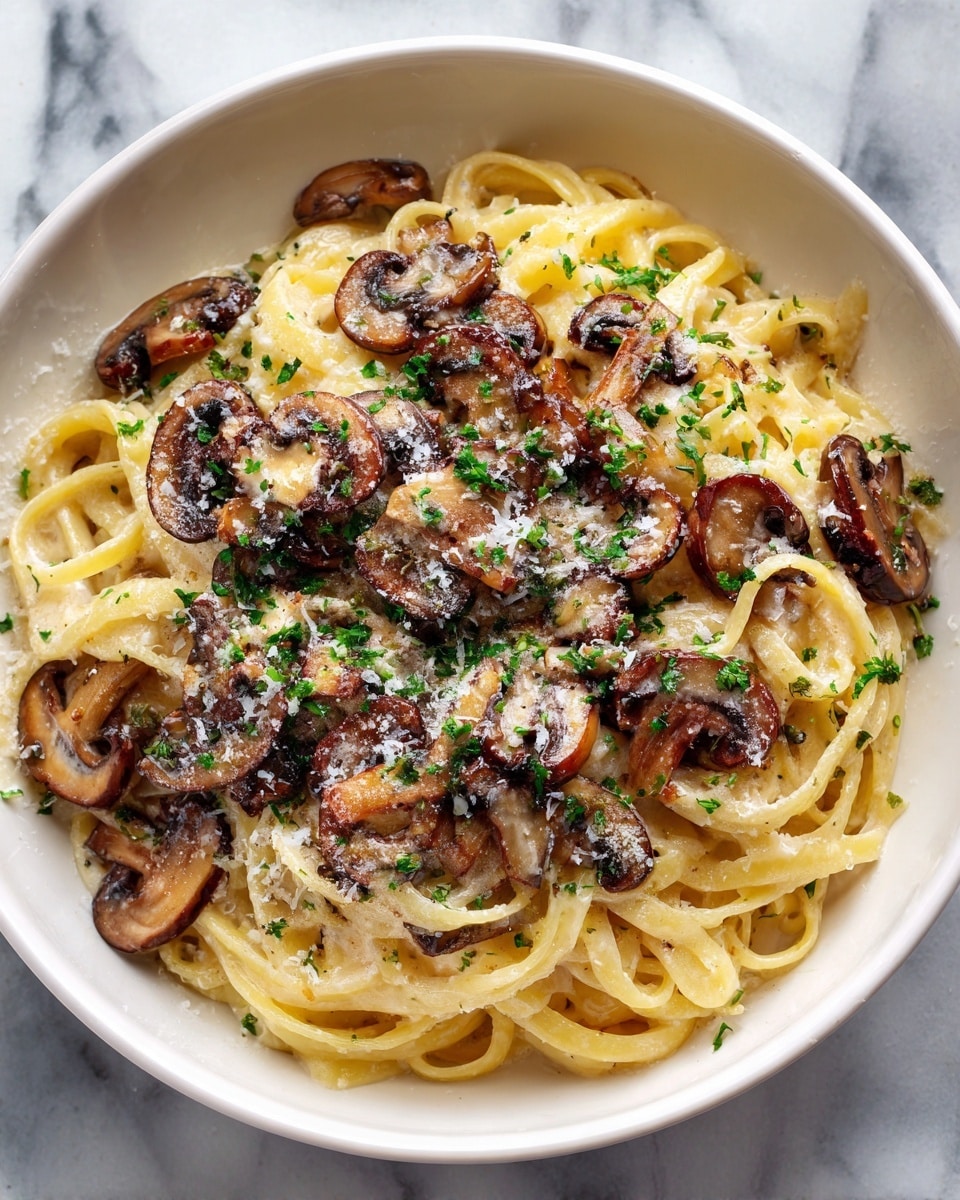 A deep white round plate holds a nest of creamy yellow fettuccine pasta coated in a smooth light sauce, layered with browned, sliced mushrooms scattered on top and around the pasta. The mushrooms have a glossy, slightly caramelized texture. The dish is sprinkled with finely chopped green herbs and a dusting of grated cheese, adding small white and green specks across the surface. The plate sits on a white marbled textured surface, lit by soft natural light, showing shadows and highlights. Photo taken with an iphone --ar 4:5 --v 7