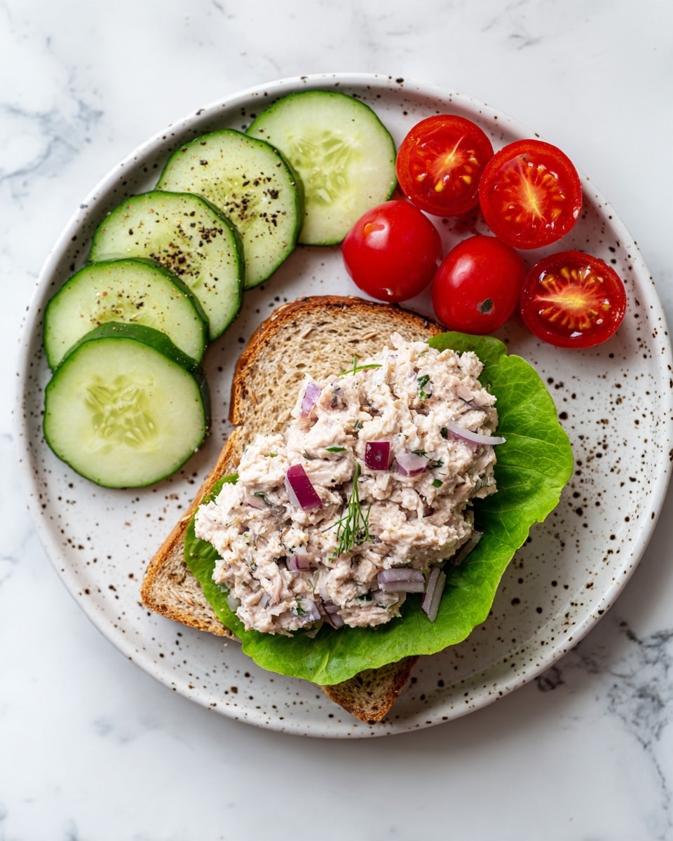 A white speckled plate holds a sandwich made with a single slice of multigrain bread as the bottom layer, topped with a large green lettuce leaf that curves slightly upward. On top of the lettuce, there is a thick, creamy tuna salad mixed with small bits of celery, red onion, and herbs, giving it a varied texture and light beige color with green and purple specks. To the left of the sandwich, there are five thin, round slices of cucumber sprinkled with black pepper, arranged in a neat line. Above the sandwich, there is a small pile of bright red cherry tomato halves, their seeds and juicy insides visible. The background is a white marbled surface. photo taken with an iphone --ar 4:5 --v 7