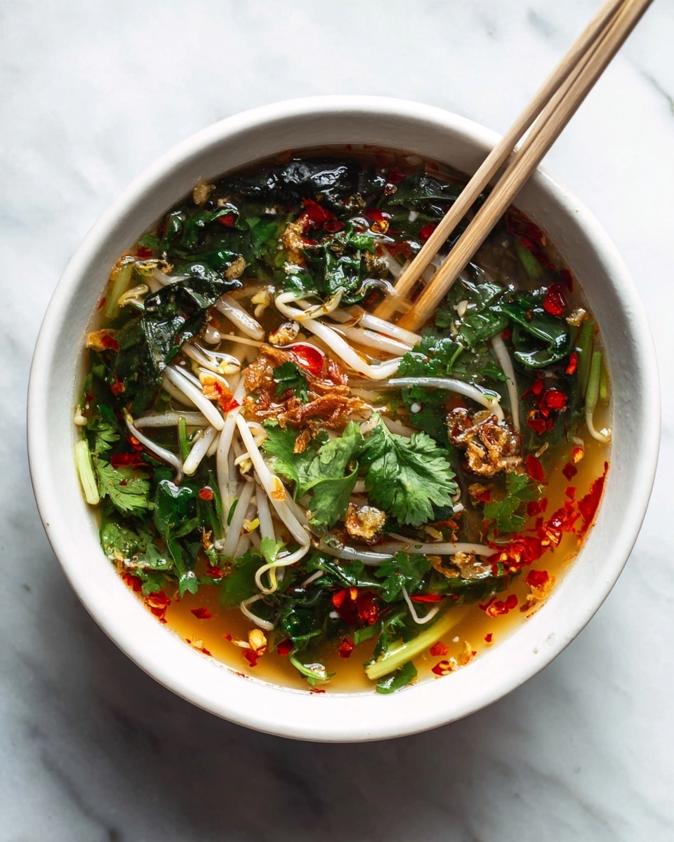 A white bowl filled with a clear broth soup with red chili oil floating on the surface, topped with bright green cilantro leaves and thin white enoki mushrooms submerged in the broth. There are bits of fried onions or garlic scattered on top, adding a crispy texture. The soup also contains dark green leafy vegetables and small pieces of red chili. A pair of light wooden chopsticks rests on the edge of the bowl. The bowl sits on a white marbled surface. Photo taken with an iphone --ar 4:5 --v 7