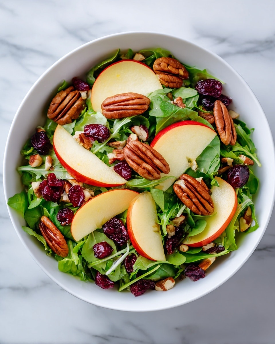 The image shows a white bowl filled with a colorful salad. The base layer is bright green arugula leaves, spread evenly. On top of the greens are slices of red and yellow apple pieces, adding a mix of red and pale yellow colors. Scattered among the apples and greens are whole pecans with a shiny brown texture. There are also small dark red dried cranberries spread throughout. Small bits of chopped nuts are sprinkled over everything, adding texture. The bowl sits on a white marbled surface. photo taken with an iphone --ar 4:5 --v 7