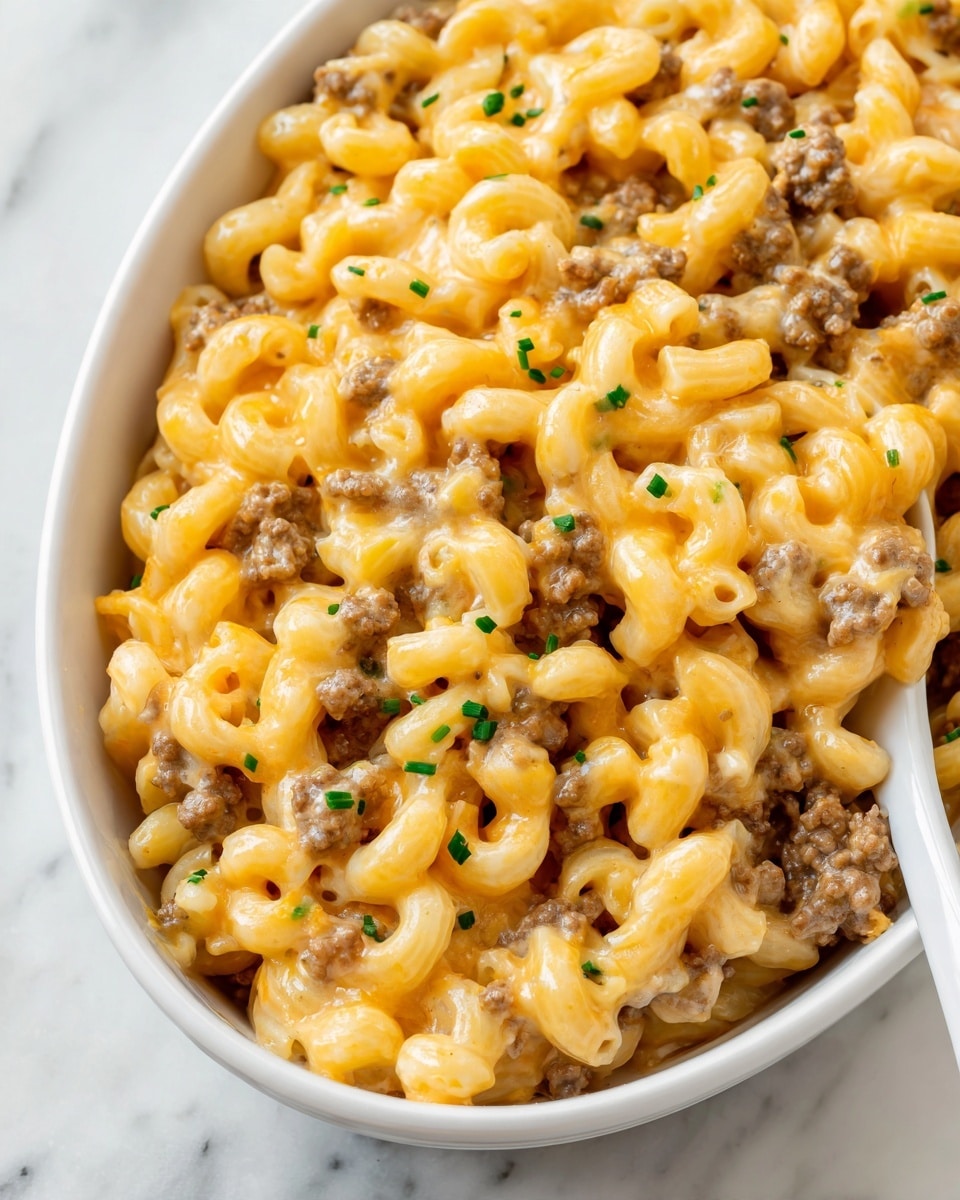 The image shows a white oval dish filled with a creamy macaroni and beef mixture. The dish has two main layers mixed together: soft yellow macaroni pasta curled in small tubes and browned ground beef coated with a rich orange cheese sauce. The sauce looks smooth and slightly shiny, wrapping around the pasta and meat. Small green chive pieces are sprinkled on top, adding bits of fresh color. A white spoon is placed inside the dish, with some macaroni and beef resting on it and the sauce visible around the edges. The dish is placed on a white marbled surface. photo taken with an iphone --ar 4:5 --v 7