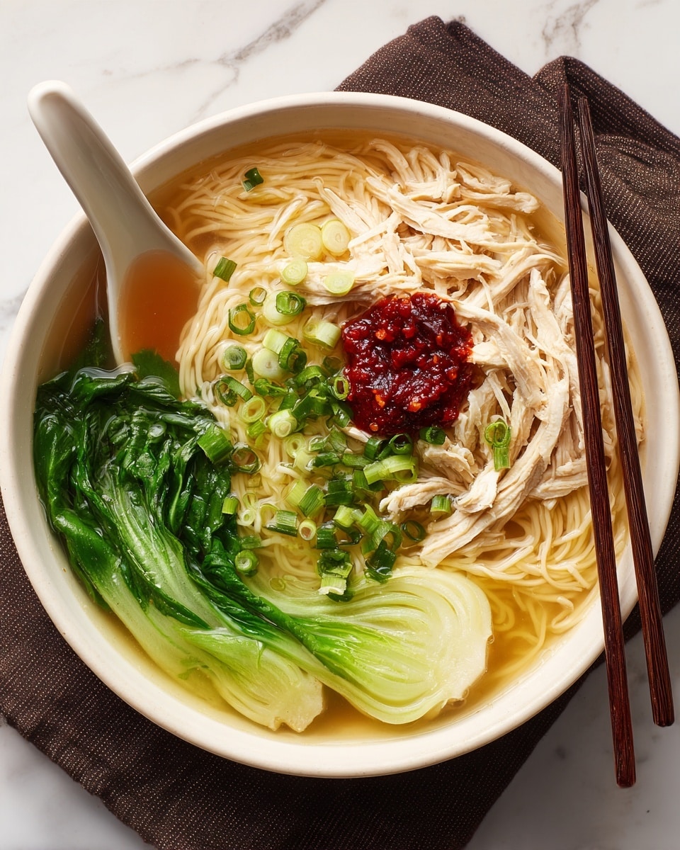 A white bowl filled with light brown broth and thin yellow noodles resting at the bottom. On top of the noodles, there are shredded pieces of pale pink chicken arranged in the center. Bright green chopped scallions are sprinkled over the chicken, along with a small dollop of red chili paste. To one side, two large, vibrant green leafy vegetables lay flat on the broth's surface. A white ceramic spoon and dark chopsticks are placed inside the bowl, resting on the sides of the noodles. The bowl sits on a soft, dark cloth on a white marbled surface with a blurred red and green jar in the background. Photo taken with an iphone --ar 4:5 --v 7