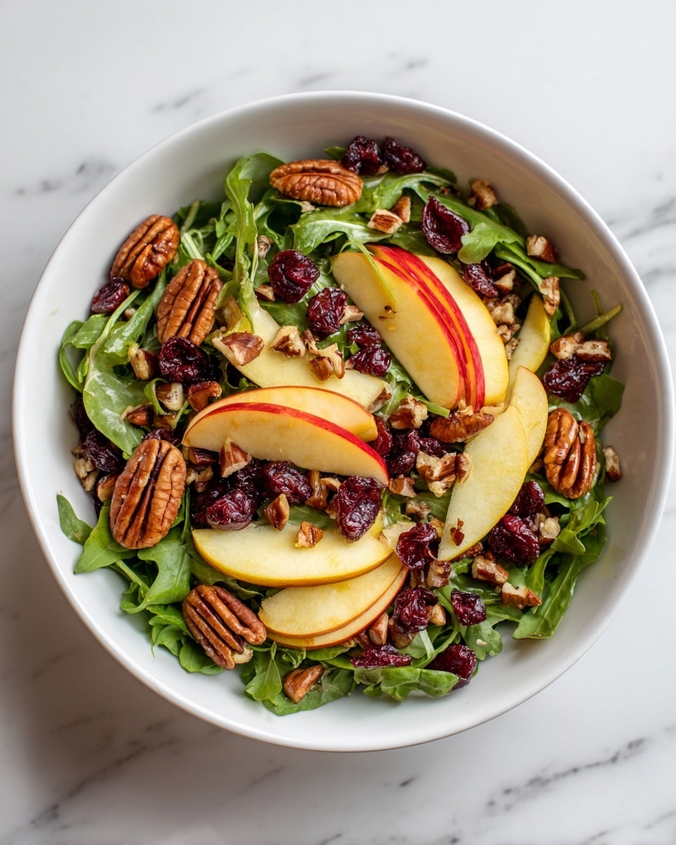 A white bowl filled with a fresh salad showing multiple layers: at the base, bright green arugula leaves create a textured leafy layer, scattered on top are thick slices of red and yellow apple wedges, deep red dried cranberries add bright spots, large shiny brown pecans spread evenly across, and small chopped nuts or seeds sprinkled all over add extra texture and detail; the salad looks colorful with a mix of red, green, yellow, and brown tones on a white marbled surface background, photo taken with an iphone --ar 4:5 --v 7