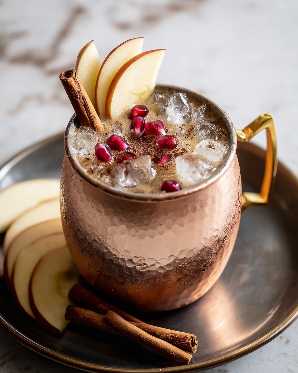 The image shows a copper cup filled with a layered drink, placed on a dark metal tray on a white marbled surface. The drink has two cinnamon sticks standing vertically on one side, a few thin slices of apple with red edges leaning against the cinnamon, and bright red pomegranate seeds scattered on top of crushed ice. The drink appears brownish, with a light sprinkle of cinnamon powder over the ice. Beside the cup, there are several apple slices leaning against it, and in the background, two similar copper cups with the same garnishes are slightly blurred. Photo taken with an iphone --ar 4:5 --v 7