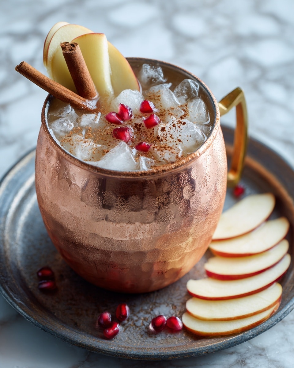 A tall copper mug filled with a brownish drink topped with crushed ice forms the base layer. On top, there are two thin, curved apple slices standing vertically, dusted lightly with cinnamon. Several bright red pomegranate seeds are scattered over the ice, adding a pop of color. Two cinnamon sticks stand upright near the apple slices on one side of the drink. The mug sits on a dark round metal tray, with a few thin apple slices placed next to the handle. The background has a soft, blurred look and the surface is white marbled. photo taken with an iphone --ar 4:5 --v 7