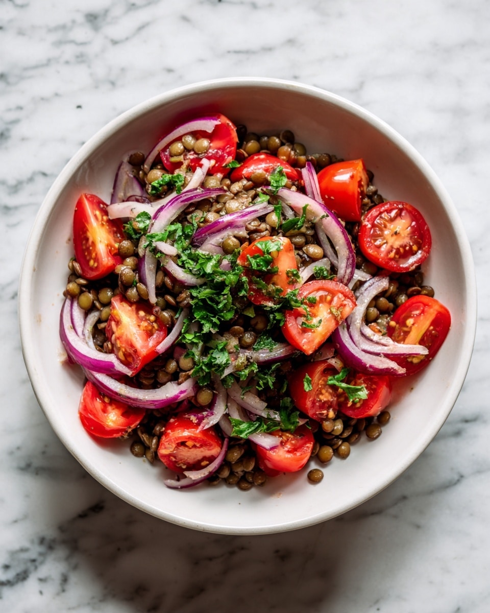 A white bowl filled with a lentil salad showing three main layers: at the bottom, soft green-brown lentils create a textured base; scattered on top are bright red cherry tomato halves with a smooth and shiny surface; thin curved slices of purple and white onion are evenly spread over the salad, mixed with small green pieces of chopped herbs giving it a fresh look. The bowl is placed on a white marbled surface. photo taken with an iphone --ar 4:5 --v 7