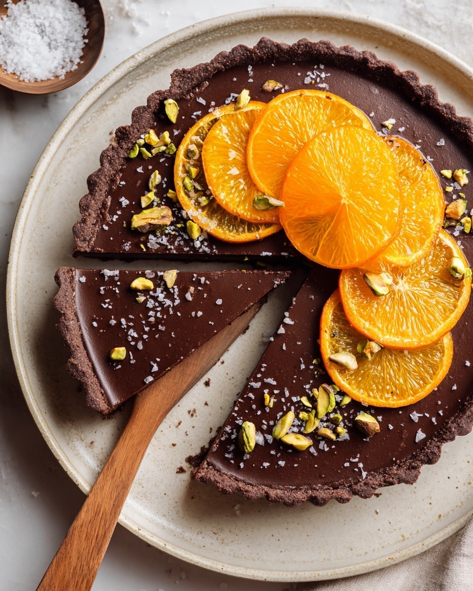 A chocolate tart with a smooth, dark brown filling and a slightly rough chocolate crust sits on a white plate. The tart is decorated on one side with thin, bright orange slices arranged in a layered fan shape, alongside small bright green chopped pistachios scattered around the edge. A single slice is cut and partially lifted by a wooden spatula, revealing the even thickness of the filling and crust. The background is a white marbled surface with small bowls holding cocoa powder and other ingredients nearby. photo taken with an iphone --ar 4:5 --v 7