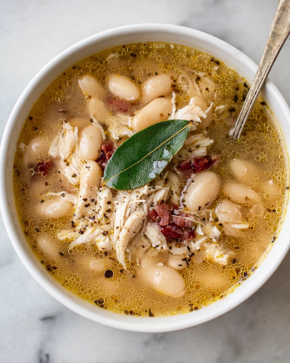 The image shows a close-up of a white bowl filled with a thick soup made of soft white beans and shredded light brown chicken pieces. There are chunks of reddish meat mixed in, and a single green bay leaf sits on top near the center. The soup broth is golden and slightly oily with small black pepper flakes floating on the surface. The texture looks smooth with the beans and meat pieces evenly spread throughout the bowl. The background is a white marbled texture. photo taken with an iphone --ar 4:5 --v 7