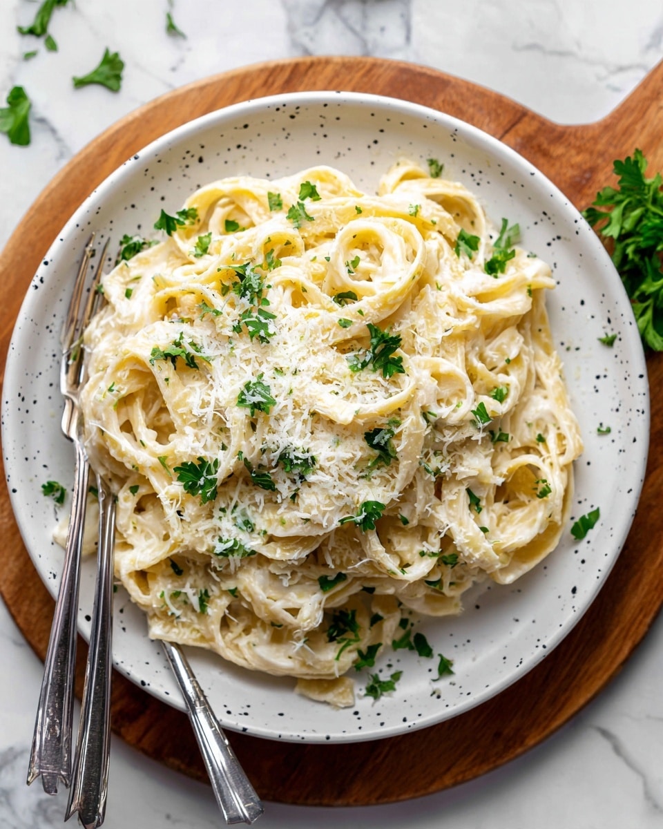 A plate of creamy fettuccine pasta with three main layers: a bottom layer of thick cream sauce, a middle layer of tender, pale yellow fettuccine ribbons coated well in the sauce, and a top layer of finely grated white cheese and small green parsley leaves scattered on top. The pasta sits on a white speckled plate, with two silver forks resting on the right side near the front. The plate is placed on a white marbled surface with some parsley leaves and cheese shavings around it. In the background, blurred green herbs sit in a ceramic container. photo taken with an iphone --ar 4:5 --v 7