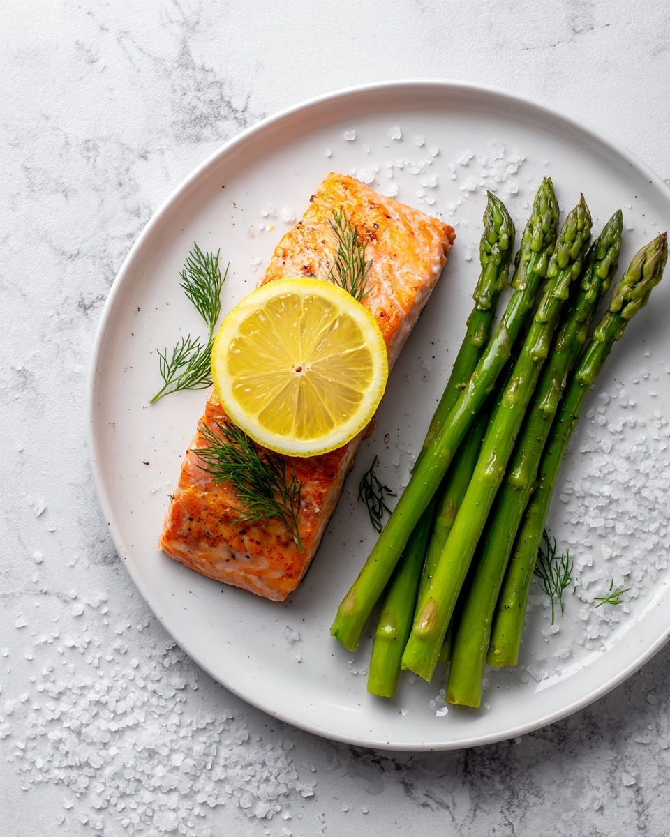 The image shows a cooked salmon fillet with a crispy orange-brown top layer on a white plate, placed on a white marbled surface. On top of the salmon, there is a bright yellow lemon slice and a small bunch of fresh green dill. To the right of the salmon, there are several long, bright green asparagus spears with a smooth texture. There are small white salt grains scattered around the plate. The photo taken with an iphone --ar 4:5 --v 7